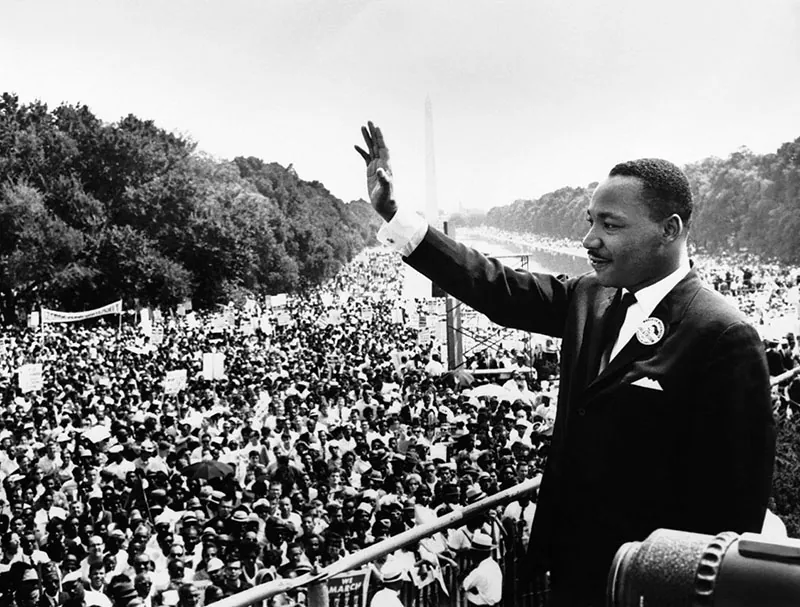 Dr. Martin Luther King, Jr. waves to an enthusiastic audience from the steps of the Lincoln Memorial with trees and the Washington Monument in the background.