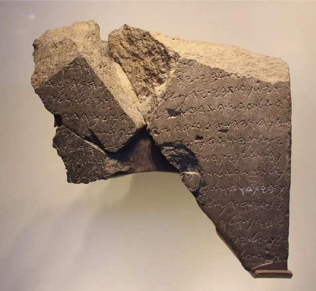 A piece of a broken brownish stone fragment is shown. Inscriptions are seen all over the stone.