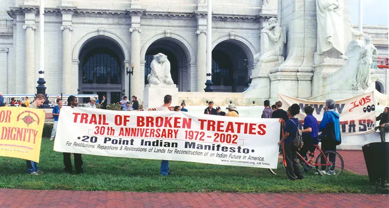 A protest in front of an official looking building with white stone pillars and a white stone lion. Prominent in the image is a banner reading “Trail of Broken Tears - 30th Anniversary 1971-2002 - 20 Point Indian Manifesto”