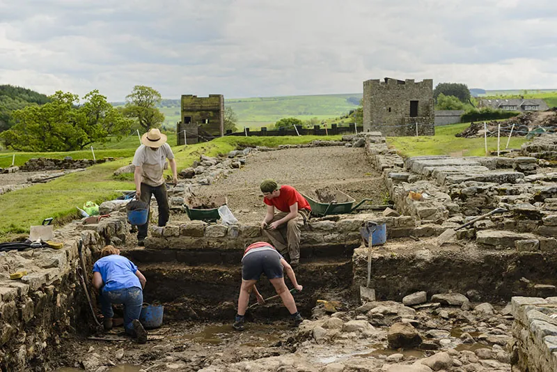 Four people are digging at an excavation site. They have partially uncovered the stone foundation and floor of an ancient building. Two square stone structures are visible in the background.