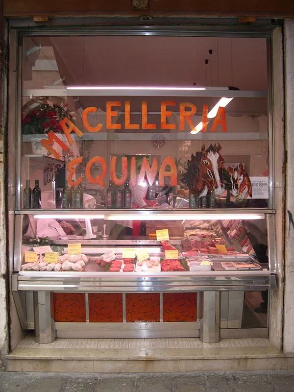 Butcher shop window with cuts of meat visible in a display case behind the glass. The sign on the window reads, Ma Celleria Equina.