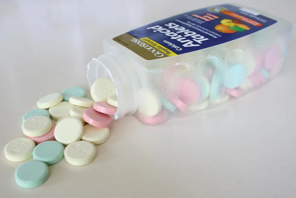 Una fotografía muestra un frasco inclinado hacia un lado con las palabras "Tabletas antiácidas" escritas en el frente. De la boca de la botella salen una serie de discos sólidos de colores.