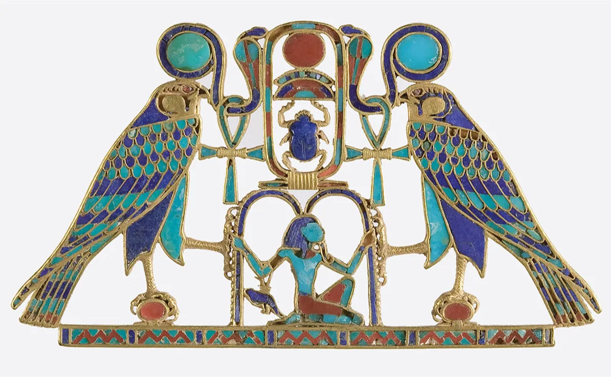 A picture of a piece of jewelry is shown. Across the bottom is a thin, long piece inlaid with red, blue, and aquamarine colored shapes (triangles and rectangles) and outlined with gold. Atop this piece in the middle an aquamarine colored person can be seen kneeling in a red waistcloth, gold beard, and blue ornamental headdress. Their arms are outstretched holding on to a carved blue and gold M-shaped arch. A blue and gold object hangs from their right arm. Above the M-shaped arch is a cartouche inlaid with red and aquamarine rectangles outlined with gold. Inside, from the top, a red circle is shown, then a colorful half circle, then an insect with blue stones and gold outlines. On either side of the cartouche are two bird-like animals in blue and aquamarine stones outlined in gold, with a snake on their heads in red, blue, and aquamarine with gold outlines, with an ankh cross hanging from each of their necks. One bird leg is standing on a red circle outlined in gold atop the long thin piece across the bottom and the other leg is grasping the M-shaped figure with the person kneeling.