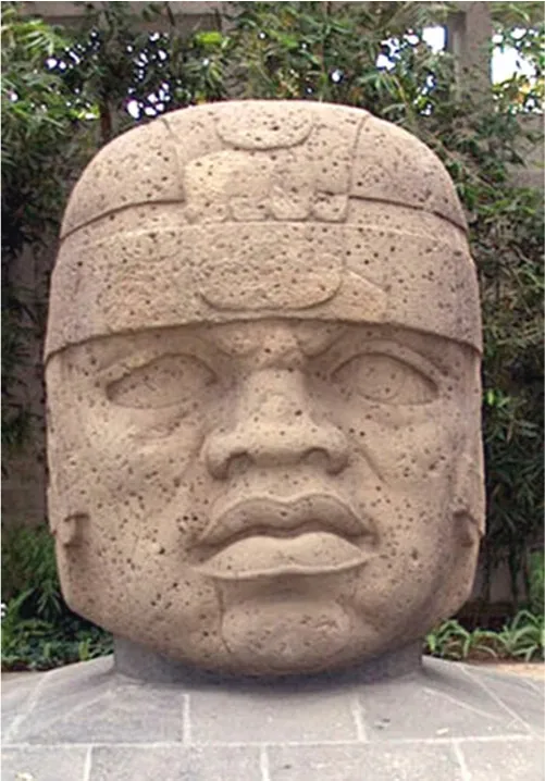 A picture of a beige stone carving of a head is shown on a gray stone pedestal with green bushes in the background. The stone shows large almond shaped eyes, a flat nose and full, parted lips. There is a helmet on his head that comes down the sides of his head. Small holes litter the stone.