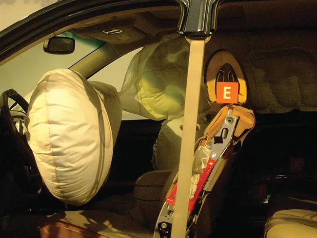 Esta fotografía muestra el interior de un automóvil desde la zona del conductor. La imagen muestra airbags inflados colocados justo delante de los asientos del conductor y del pasajero y a lo largo del lado del pasajero sobre las ventanillas. Un gran airbag redondo cubre el volante.