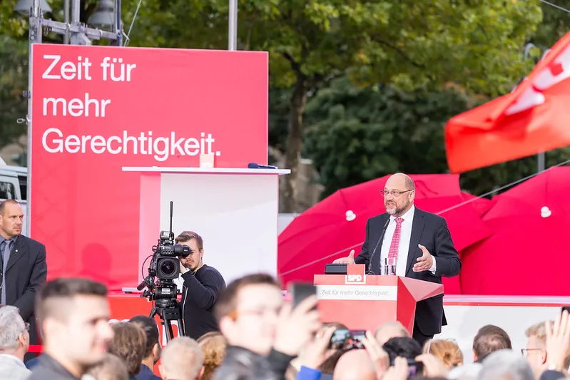German politician Martin Schulz stands at a red podium on an outdoor stage speaking to an assembled crowd while a videographer films the scene.