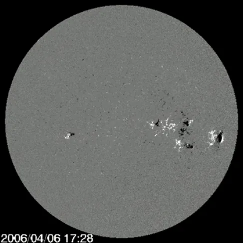 A magnetogram of the sun, which appears as a gray disc against a black background, with white and black spots scattered on it. Most of the spots are concentrated in the center right part of the image.