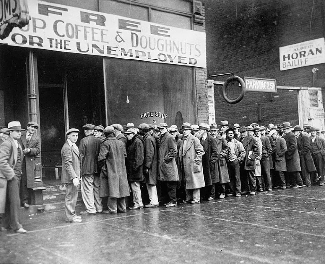 Men are lined up outside a building that has a sign that reads “Free Cup Coffee and Doughnuts for the Unemployed.”