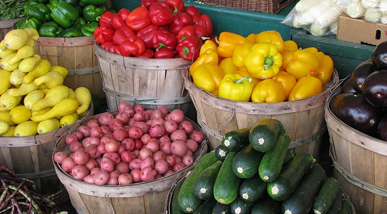 Na fotografii przedstawione są koszyki z różnymi warzywami (cukinia, papryka ziemniaki i inne) stojące na targu.