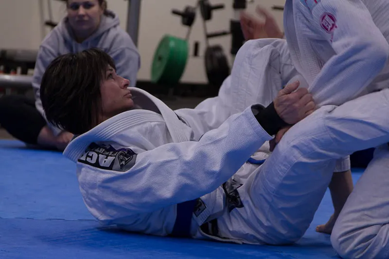 Two people in jiu-jitsu uniforms sparring during a jiu-jitsu training session as a woman in the background watches them.