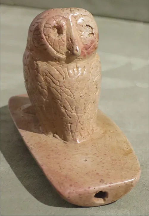 A carving of an owl in speckled, pale, brown marble is shown. It is perched on a smooth, half circle shaped long rectangular piece with a small hole in the front made out of the same marble. The owl shows large deep inset eyes, a long thin pointy beak and feathers carved into the head and body. The object lies on a gray surface and a shadow can be seen underneath.