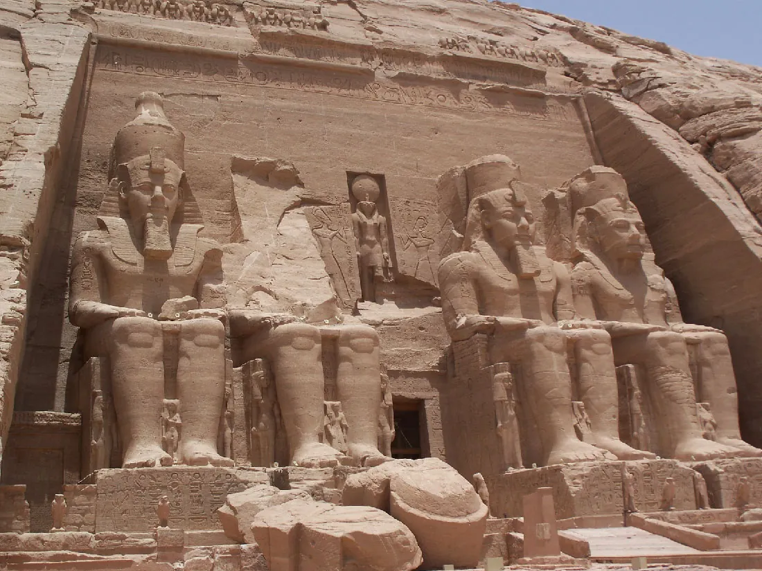 A picture of sand colored statues against a hill are shown. Four figures are shown in a row, two on each side, sitting on thrones with footrests. The pieces of the upper half of the second figure are seen on the ground in pieces in front of the feet of the statue. The other three figures wear tall headdresses with wide sides that extend down to their shoulders and chests. They are shown with long beards, decorative arm braces and both hands resting in their laps. All four are barefoot. A small statue is shown carved into the wall between the heads of the large statues. Two female figures are seen outlined simply on either side of the inset statue. Below the inset statue is an opening. Along the footrests of the large statues are hieroglyphics carved into the stone. Bird statues are seen in the forefront around the statues. The top of the carving has symbols and script carved into the stone as well as figures above that. The sides are the rocky hill.