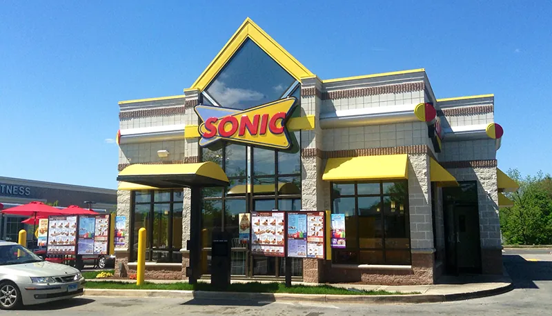 A Sonic drive thru is shown. The Sonic logo appears on the top center of the building; menus are posted near the curbs of the drive thru lane.