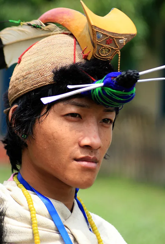 A color photograph of a the head and shoulders of young man with tan skin and black hair. He wears an elaborate hat consisting of a basket-like base topped by a feather and a thick, rolled decoration. The front of his hair is secured into a hank against his forehead, wrapped in a length of fabric and pierced by two long sticks.