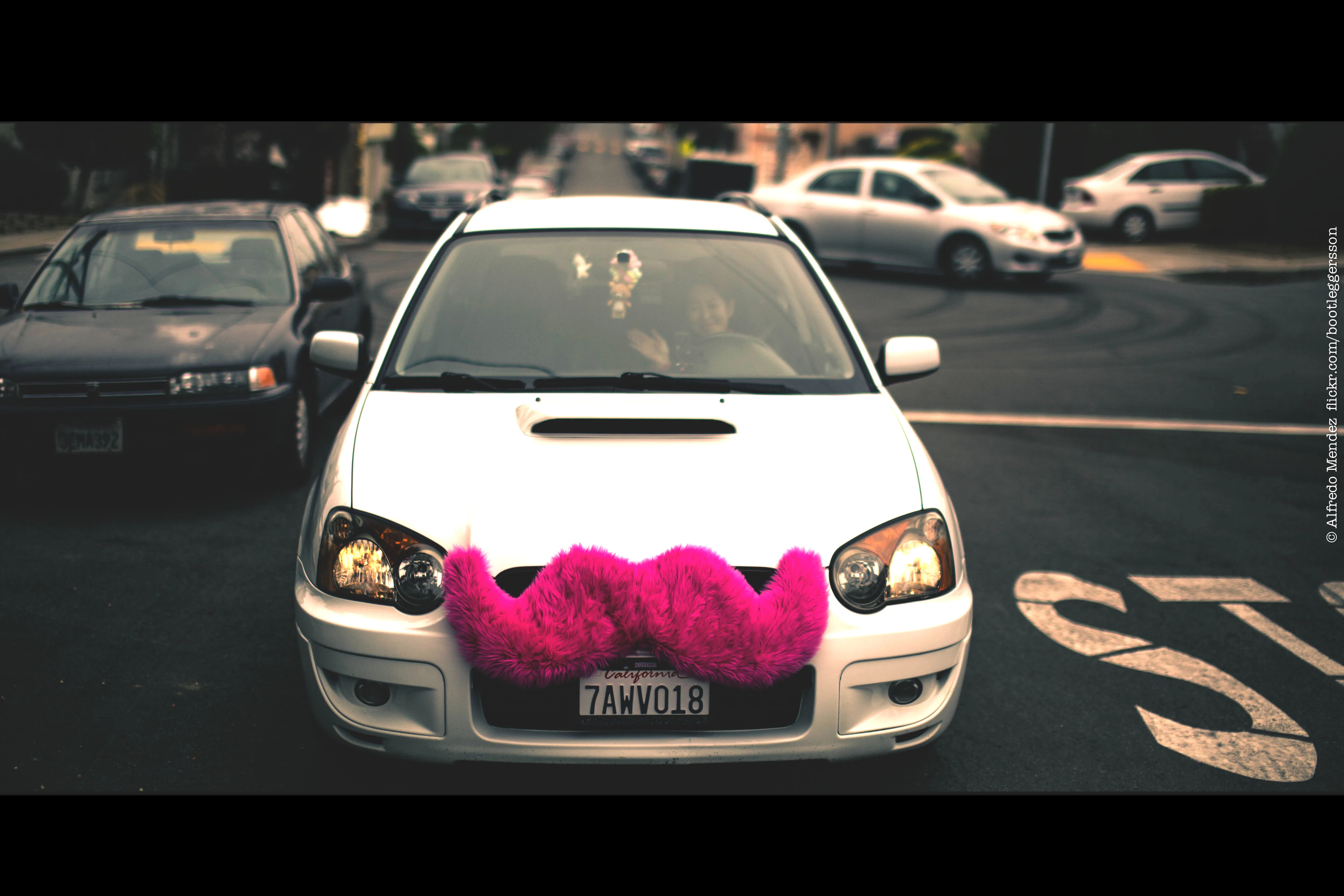 A photograph shows a car with a large, fuzzy, pink moustache attached to the front grill of the car.