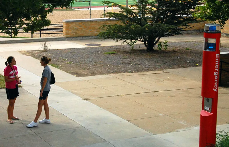 A photo shows two female college students talking to each other on a college campus. A large emergency phone with buttons and siren appears toward the right.
