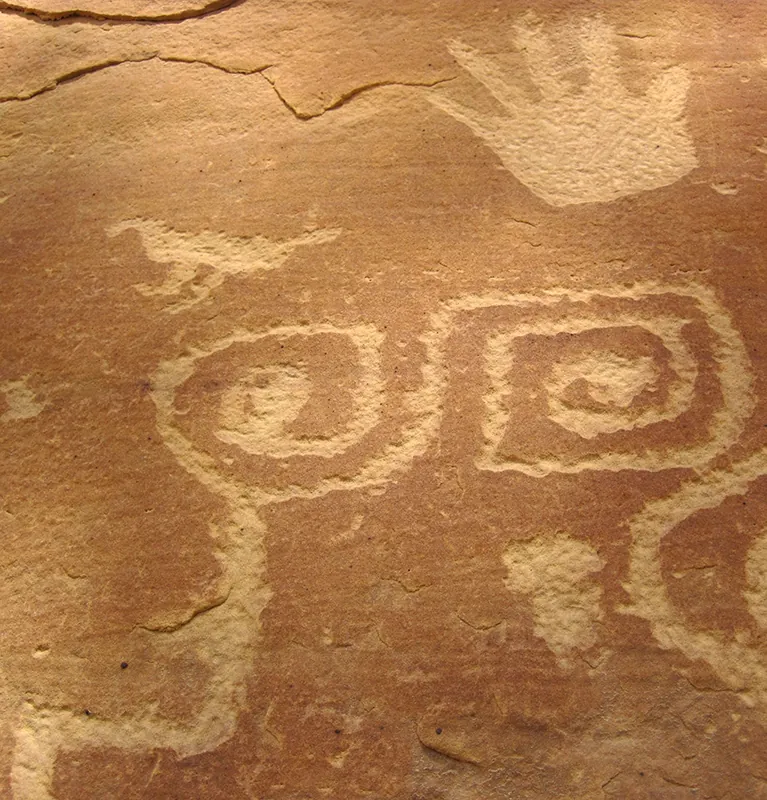 Cliff face displaying designs created by carving out a portion of the surface, revealing lighter-colored rock beneath. Designs include two connected spiral shapes, a hand, and a bird.