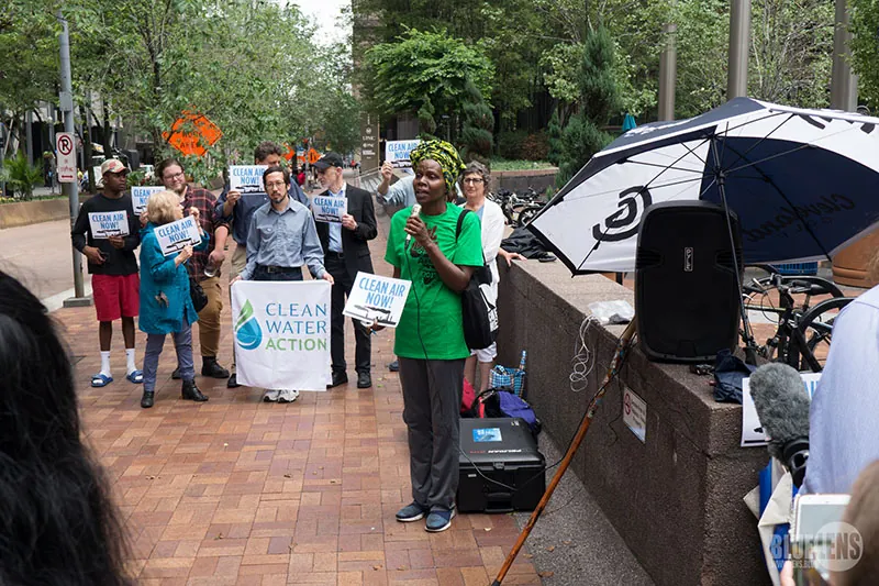 A group of people holding a rally in a public space. They hold signs reading “Clean Air Now” and “Clean Water Now.” One woman is holding a microphone and speaking.