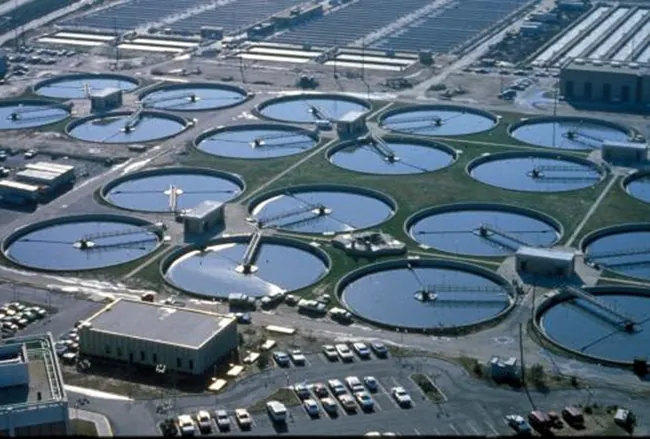 Se muestra una fotografía en color de una instalación de tratamiento de aguas residuales de gran volumen. En el centro de la fotografía se ven diecinueve piscinas grandes circulares de agua en tratamiento. En primer plano se ve un edificio y un estacionamiento.