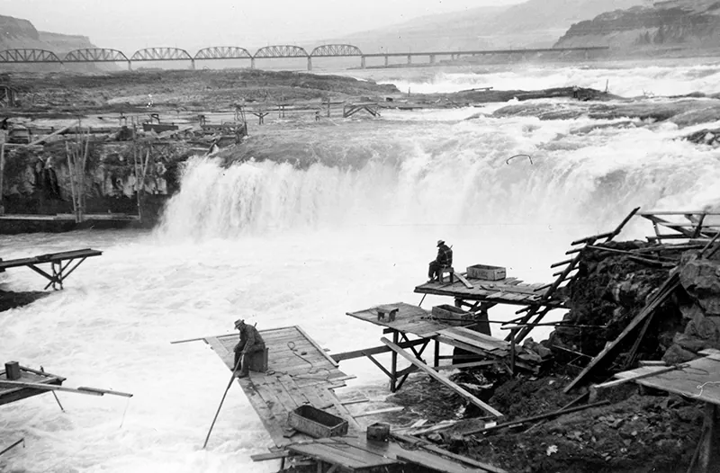 Two men sit on wooden platforms on the bank of a river, holding long poles with the ends submerged in the water. A waterfall is visible behind them.