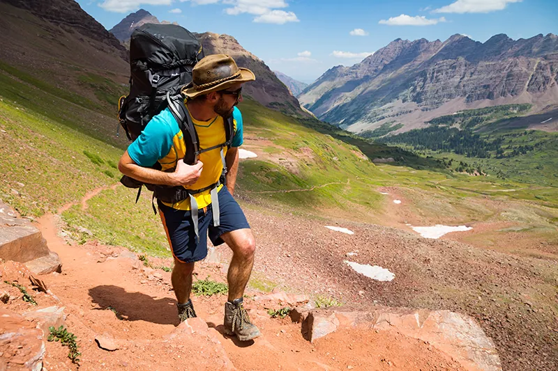 A hiker stands in front of a large mountain range. The person is carrying a large hiking pack on their shoulders and is wearing hiking boots.