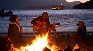 A man plays the guitar while his friends sing along around a campfire.