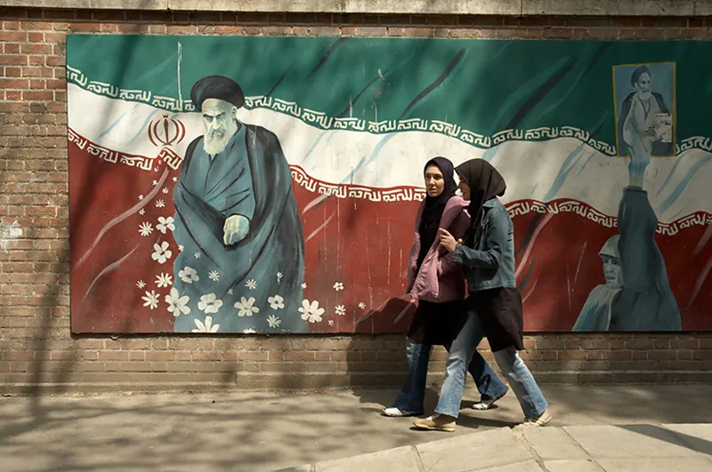 Two people wearing head scarves walk down the street in front of an Ayatollah Khomeini mural hung on the side of a brick building.