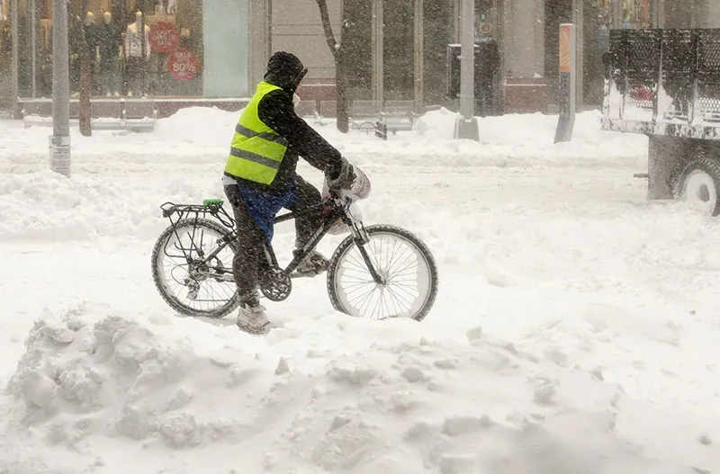 A bicycle messenger or delivery person stands with their bike still on a snowy street while a truck drives in front of them.