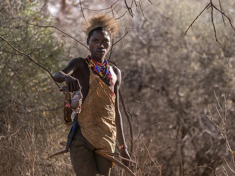 A Hadza man standing in a brushy area holding a small animal he killed in a hunt.