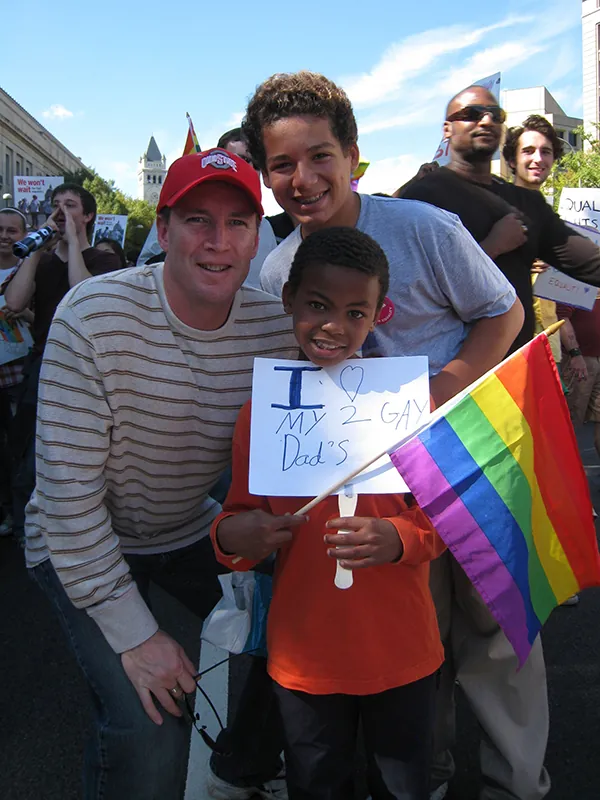 A White man, a teen boy and a young Black boy areat a gathering posing for a picture. The Black boy is holding a sign that reads I heart my two dads. He is also holding a rainbow flag.