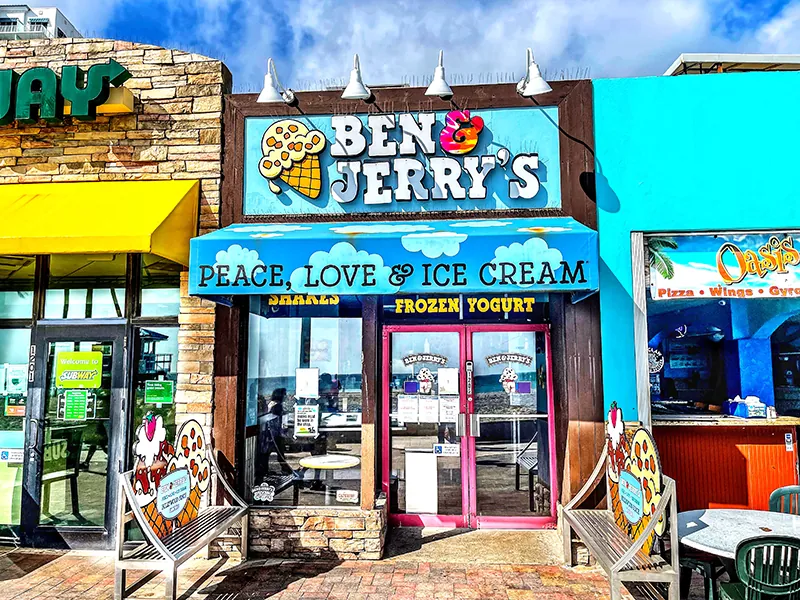 A Ben & Jerry’s storefront is between other stores. An awning has clouds and sky painted on it with the words Peace, Love & Ice Cream.
