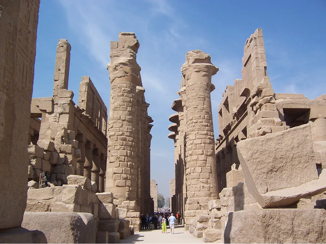 A picture of a stone building in ruins is shown. Two rows of five tall sandy-colored stone columns stand in a row in the middle. Pieces are missing at the top and parts of the columns are missing bricks. In between the columns a large group of people can be seen standing and walking in various types of clothing. On both sides of the columns a two-tiered building can be seen in ruins. Wall and roofs are missing from the top tiers and large openings with tall decorated columns can be seen on the bottom tiers and pieces on the second tiers. In the forefront of the picture on both sides large pieces of sandy-colored stone can be seen laying on the ground, some atop each other. The ground is flat and sandy-colored and a blue sky with pale white clouds can be seen in the background.