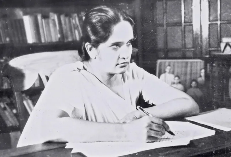 Sirimavo Bandaranaike sits at a desk holding a pen over a small stack of papers.