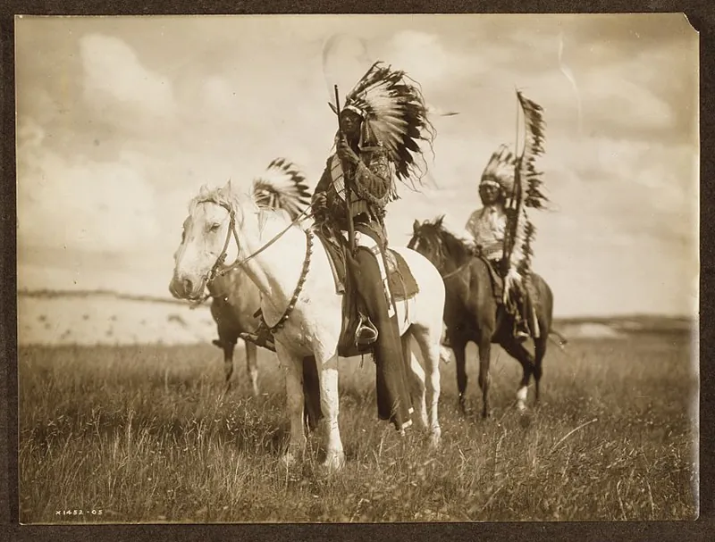 Three Native American men pose on horses on a grassy plain. They wear large feather headdresses and hold staffs and bows also decorated with feathers.
