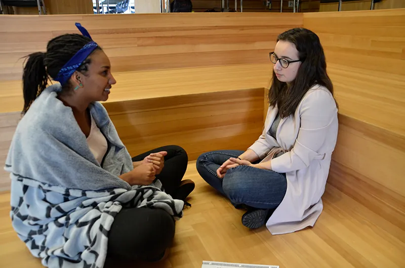 A photo shows two young women sitting on the floor of a wooden cubicle and talking to each other.