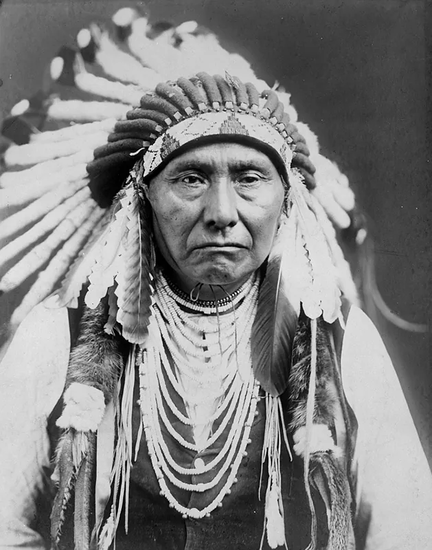 Native American man wearing a large feather headdress and many strings of beads, staring straight ahead with a serious expression.