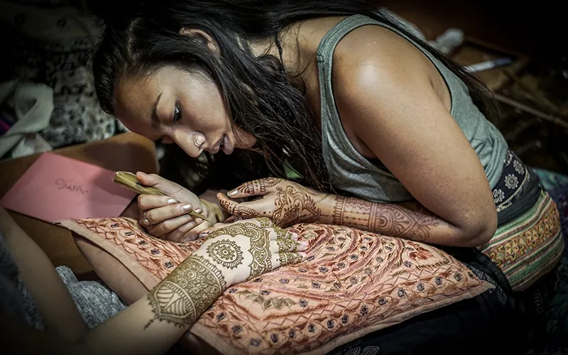 A woman with hands and forearms painted with henna art painting another woman’s hands and forearms.