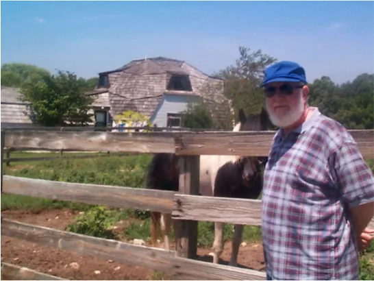 A color photograph of a man in a plaid shirt posing in front of a geodesic dome. He is tall, with a white beard, and stands with his arms held behind his back. The dome is large, with windows appearing at two different levels. The overall shape is that of a half circle, although the dome is composed of various flat pieces fitted together. A horse stands beside the man.