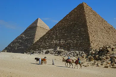 Obraz przedstawia dwóch ludzi jadących na dwóch wielbłądach i dwukołową bryczkę zaprzęgniętą w konia przed dwiema piramidami w Egipcie