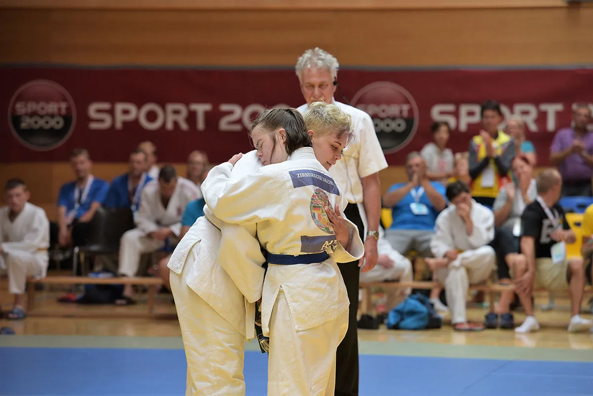 Two people wearing judo uniforms and rank belts embrace at the center of a mat while a referee and audience look on.