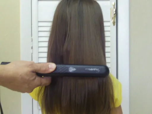 A hair straightener is being used on a girl’s long dark hair. A large section of the hair is sandwiched between the two flat irons of the hair straightener, which is moved down the hair, applying heat.