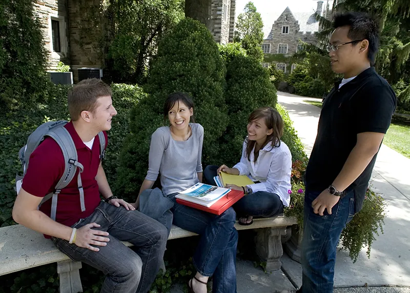 A group of students sitting outside, speaking, and smiling. Several hold books and one wears a backpack.