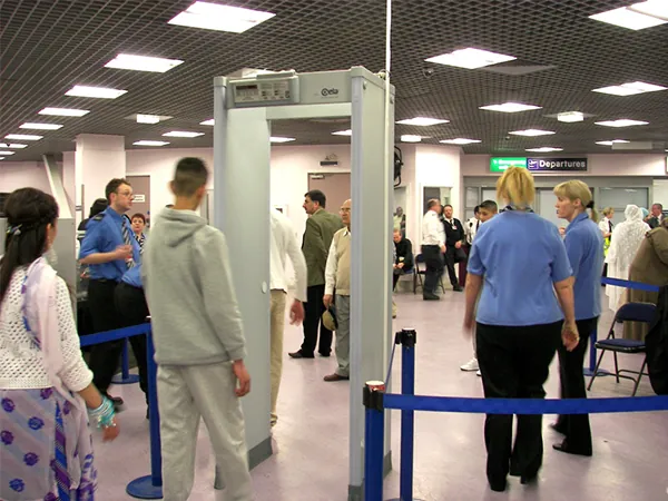 Zdjęcie przedstawia ludzi ustawionych w kolejce do bramki detektora z wykrywaczem metali na lotnisku. 