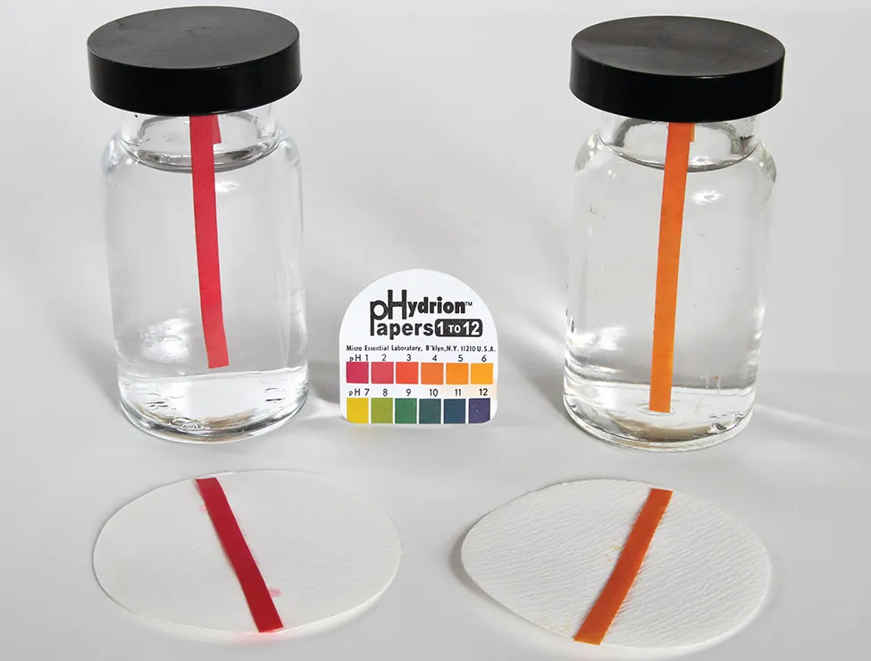 Esta imagen muestra dos frascos que contienen soluciones transparentes e incoloras. Cada frasco contiene una sola tira indicadora de p H. La tira en la botella de la izquierda es roja, y una tira roja similar se coloca en un círculo de papel de filtro delante de la botella en la superficie sobre la que se apoyan las botellas. Del mismo modo, la segunda botella de la derecha contiene una tira de color naranja y se coloca una tira de color naranja delante de ella en un círculo de papel de filtro. Entre las dos botellas hay un paquete de papeles p Hydrion con una escala de colores p H en su cubierta.