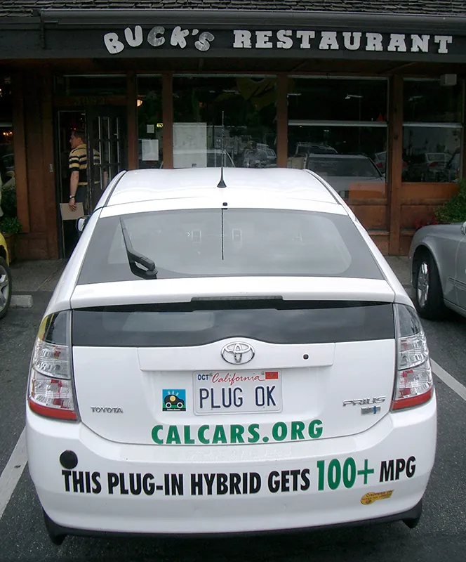 A Toyota is parked in a parking space. The license plate reads Plug OK. Beneath the license plate it says Calcars.org. On the bumper is written “This Plug In Hybrid gets 100+ M P G”.