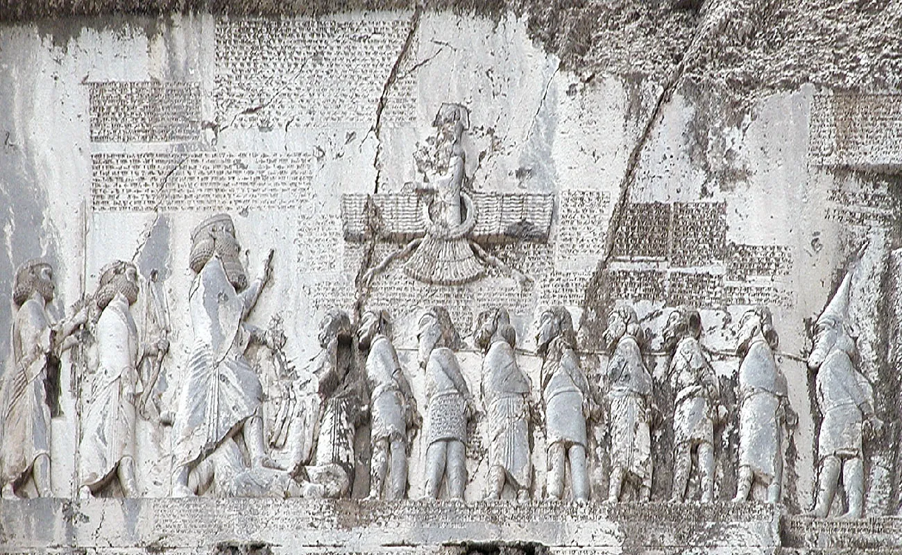 A picture of a wall carving is shown. It is worn, chipped, pieces are missing, and cracks are visible in many places. Across the lower portion, two men are shown on the left wearing long robes, holding a spear and a bow, with long hair and long beards. They face to the right. In front of them stands a larger man in a long robe facing to the right, crown on his head, with long hair and a long beard. He is holding his right hand up, holds a bow is his left hand, and his right foot is resting atop a body lying on the ground facing him. Nine shorter figures are shown to the right standing in a line facing left with their hands behind their backs and ropes connecting their necks. They wear a variety of shirts, robes, and cloths around their waists. All have beards and one man at the end wears a tall pointed hat. A figure is shown floating above in a long ruffled robe inside a circle with some decorative lines on the sides. On the wall above the figures there are columns of worn out writing as well as across the bottom under the figures.