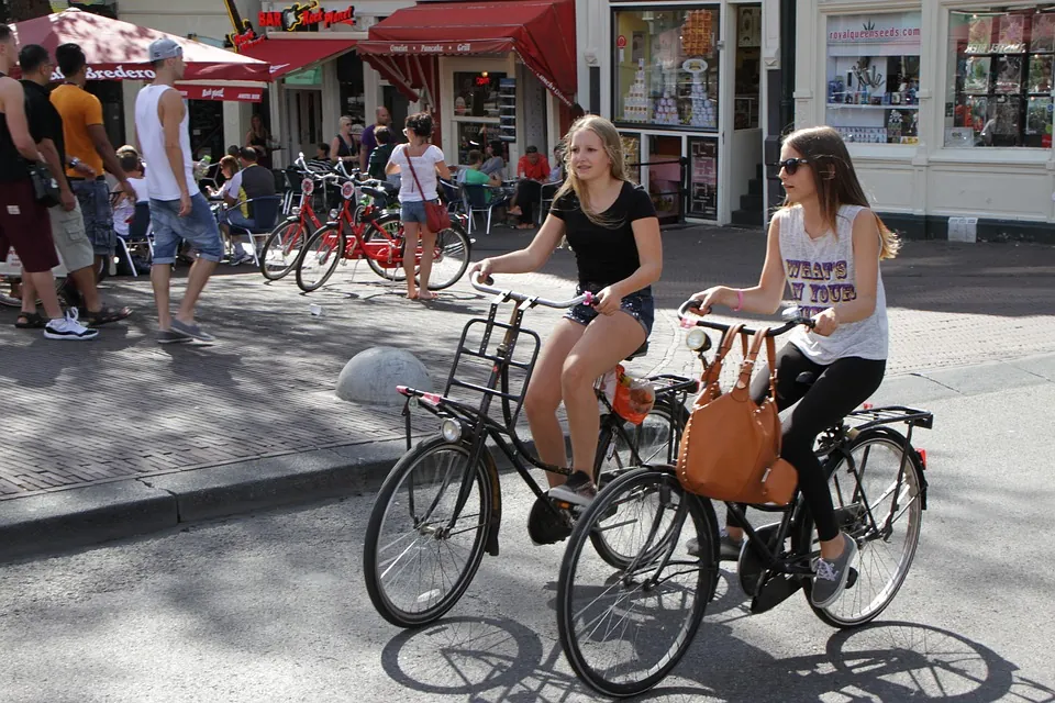 Zdjęcie pokazuje dwie dziewczyny jadące na rowerach ulicą obok zabudowań.