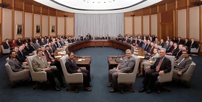 A group of professionals wearing suits sit in semi-circle rows in a board room.