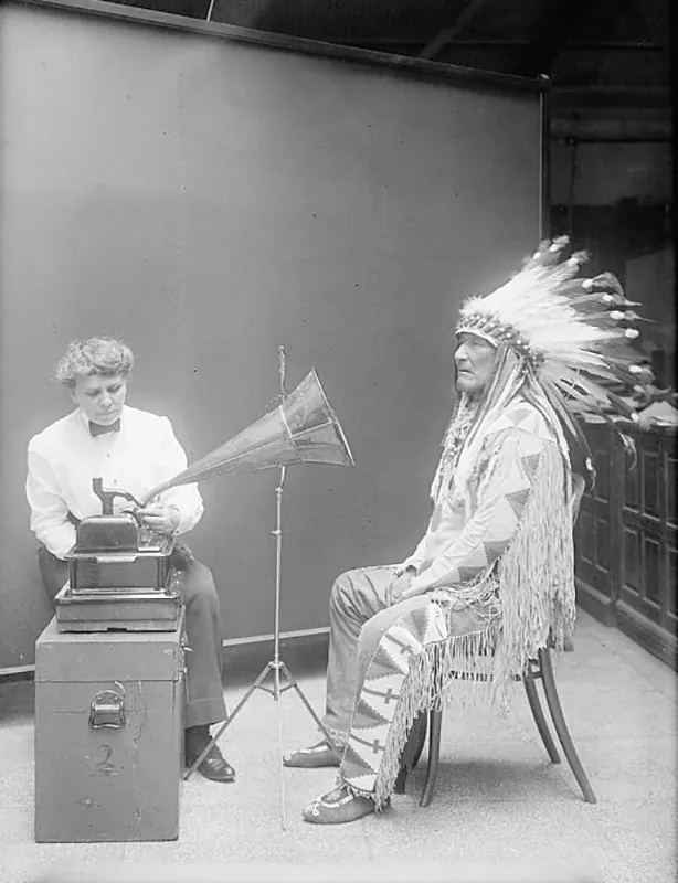A woman wearing a dress coat and pants sits behind a phonograph. A Native American man wearing a full headdress sits in front of the phonograph’s speaker.