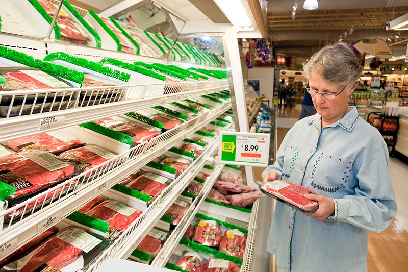 A shopper stands in front of the meat section of the grocery store, holding a package of beef.